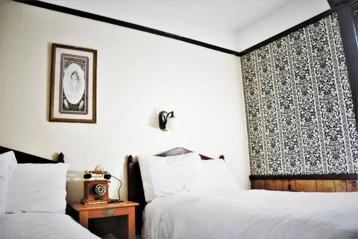 A hotel room with a bed, a nightstand with a telephone, a framed picture on the wall, and patterned curtains.