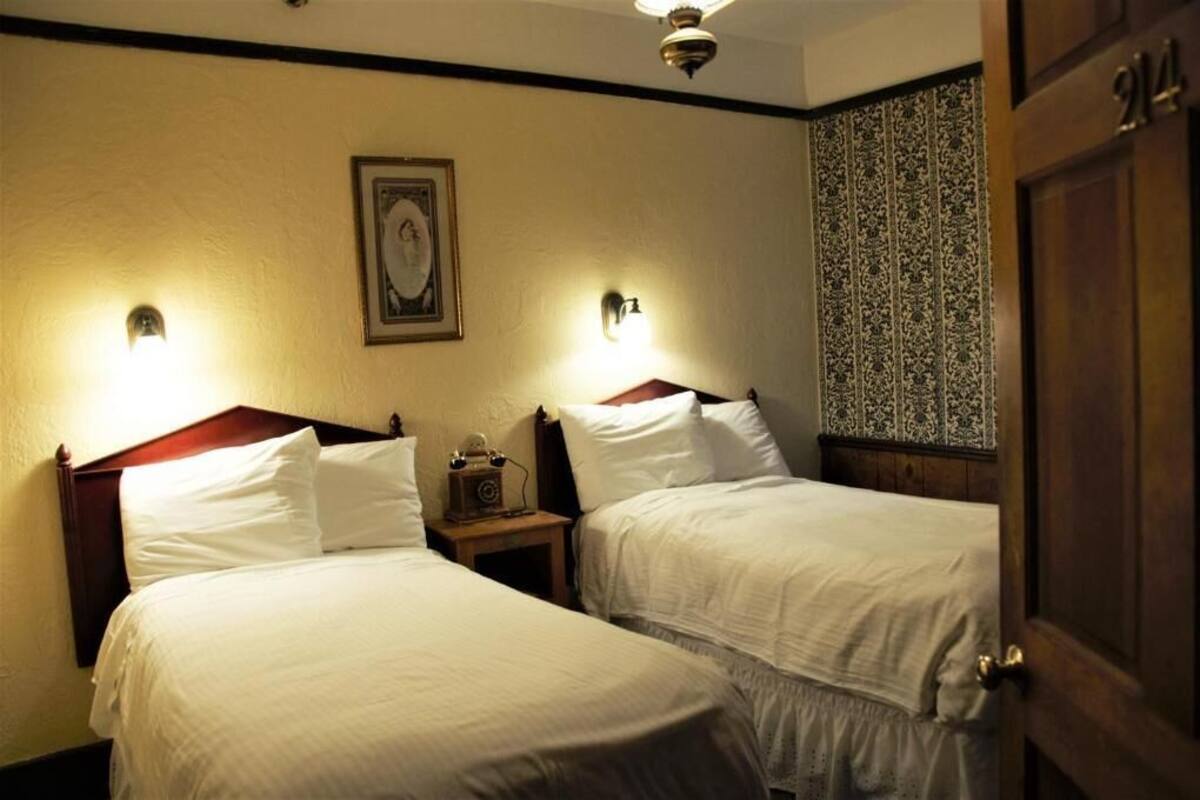 A hotel room with two beds, a nightstand with a radio, and a door with a number.
