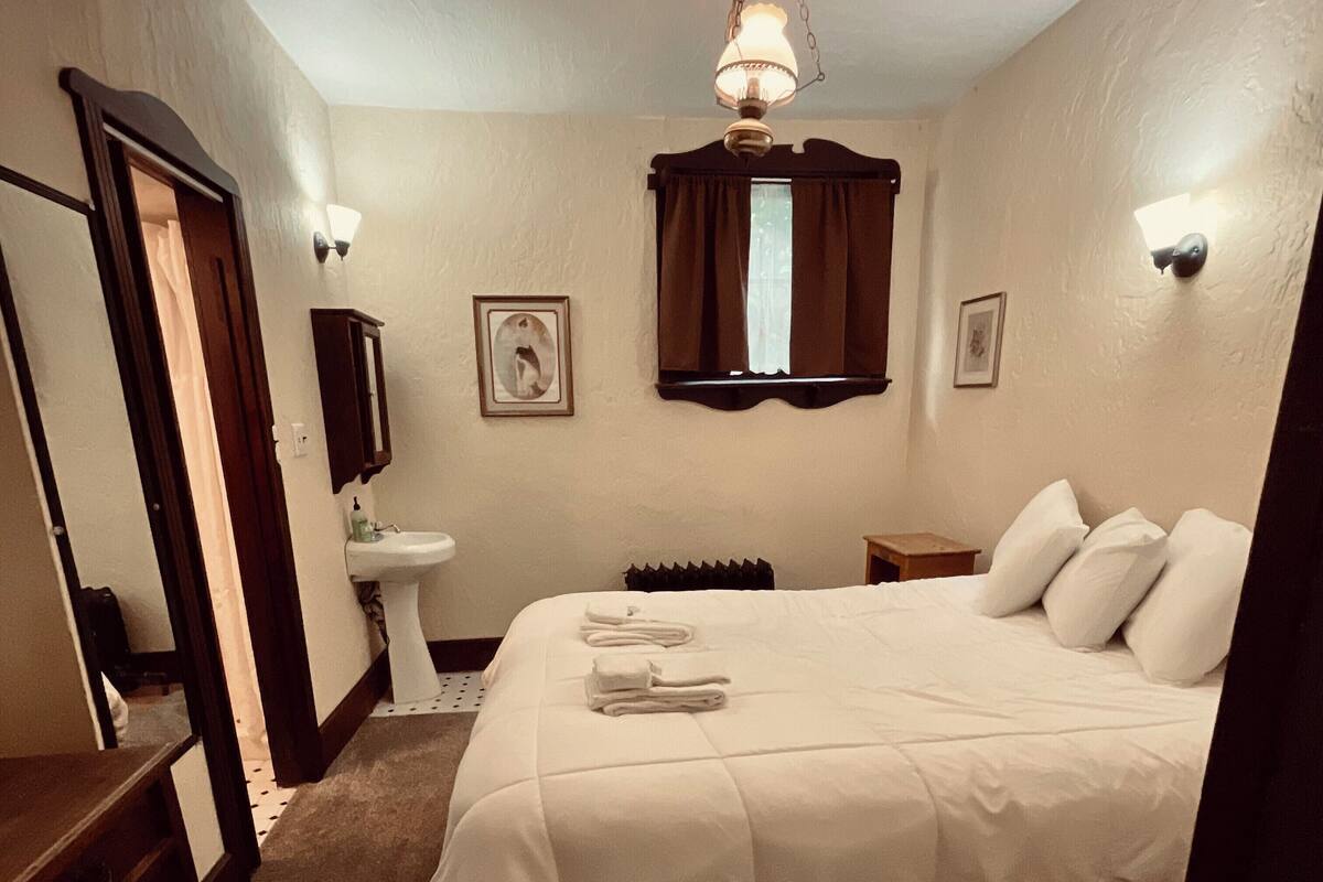 A hotel room with a large bed, a bedside table, a nightstand, a window with curtains, a wall-mounted light fixture, and a framed picture.