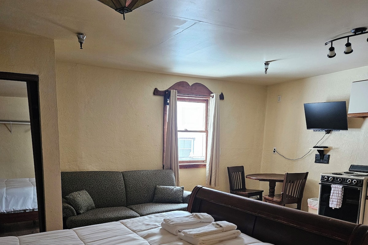A hotel room with a large bed, a sofa, a dining table, a TV, and a stove.