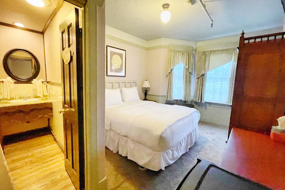 A hotel room with a bed, a bedside table, a mirror, and a window with curtains.