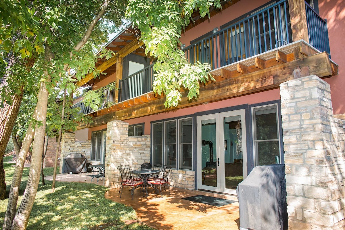 A two-story house with a stone exterior, a covered patio with a table and chairs, and a large window overlooking the yard.