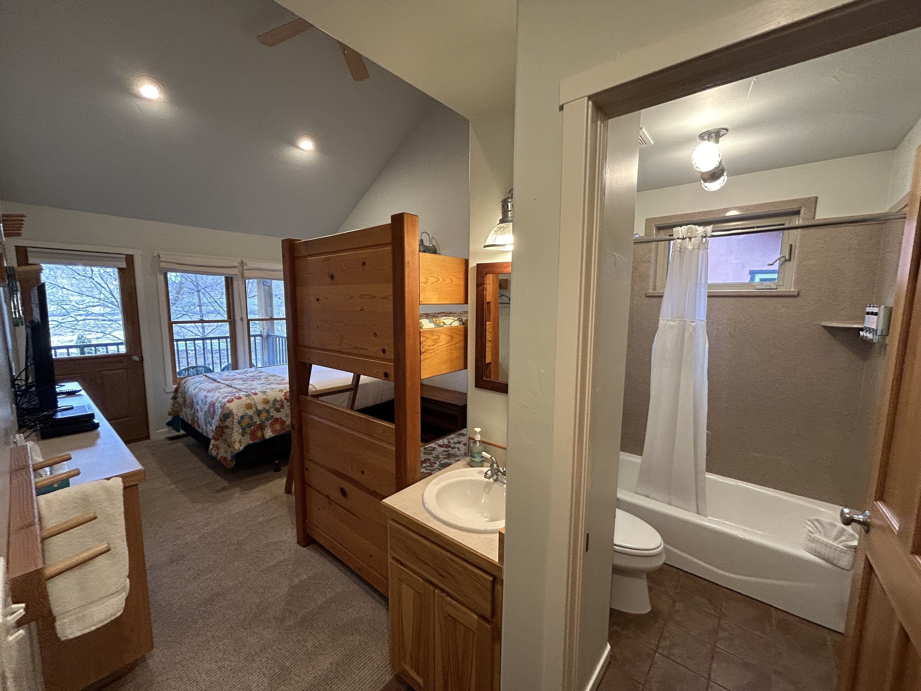 A bedroom with a wooden bunk bed, a desk with a computer, a large window, and a bathroom with a bathtub and toilet.