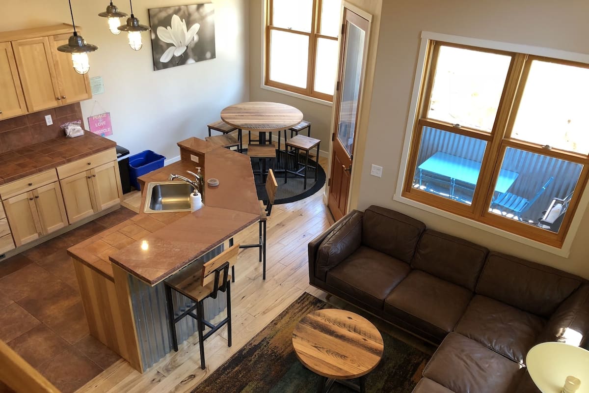 A compact kitchen with a central island, a dining area with a round table, a brown leather sofa, and large windows offering views of the outside.