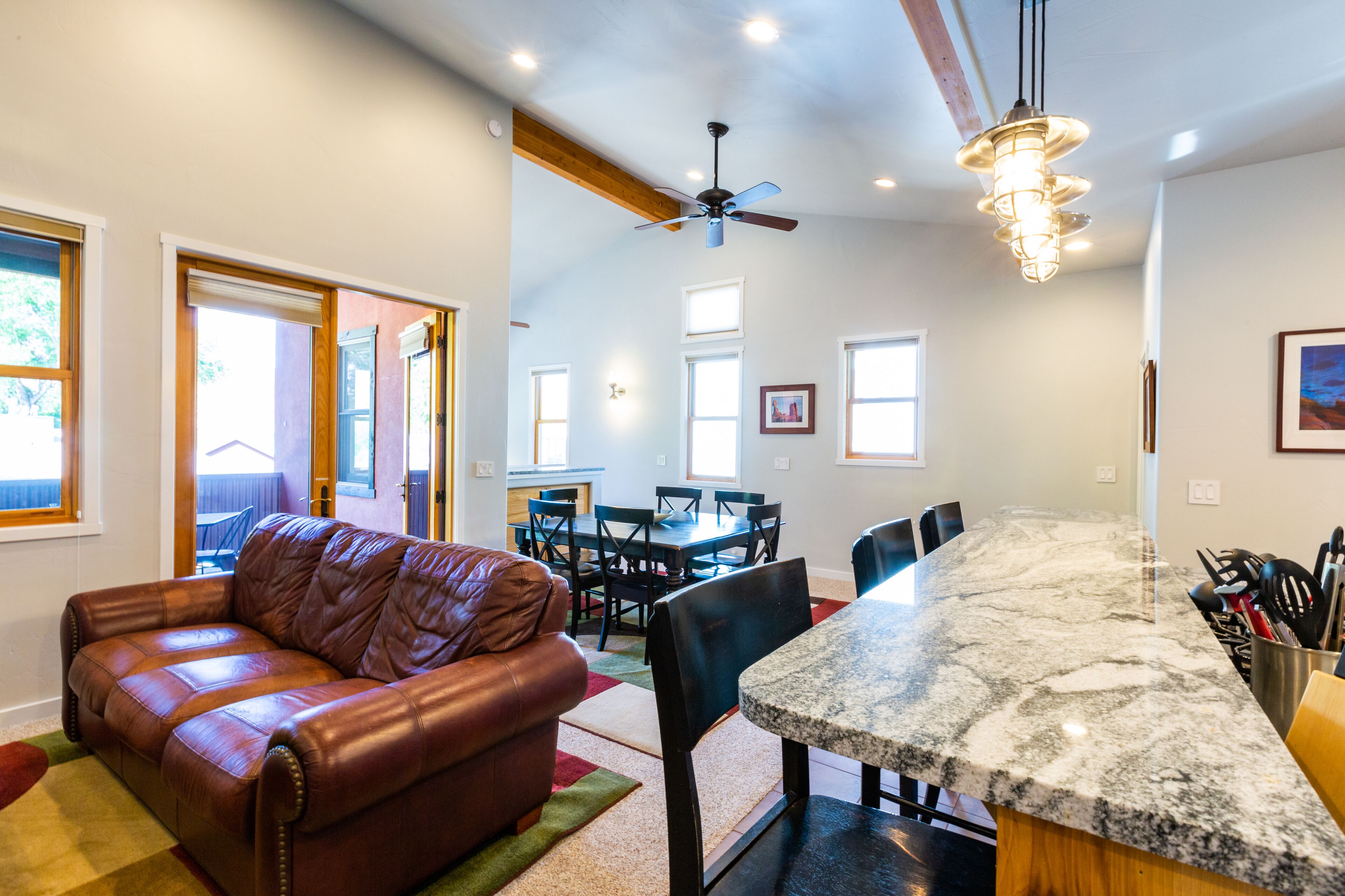 A spacious living area with a leather sofa, a dining table with chairs, a kitchen island with a granite countertop, and a ceiling fan.
