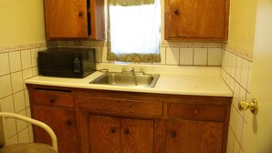A small kitchen with wooden cabinets, a microwave, a sink, and a window with a curtain. - Sahara Motel (Anaheim)