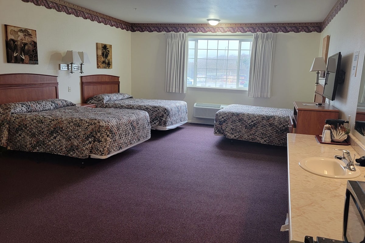 A hotel room with three beds, a large window, a bathroom with a sink and mirror, and a television.
