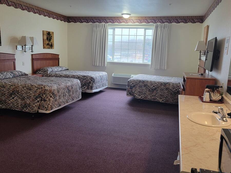 Habitación de hotel con tres camas, un ventanal grande, un baño con lavamanos y espejo, y un televisor.