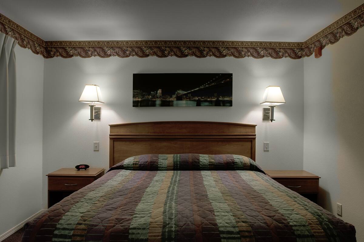 A neatly made bed with a patterned quilt, two bedside tables with lamps, and a cityscape painting on the wall.