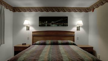 Een netjes opgemaakt bed met een quilt met patroon, twee nachtkastjes met lampen en een stadslandschap schilderij aan de muur.