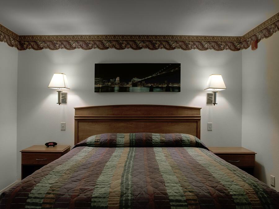 Een netjes opgemaakt bed met een quilt met patroon, twee nachtkastjes met lampen en een stadslandschap schilderij aan de muur.