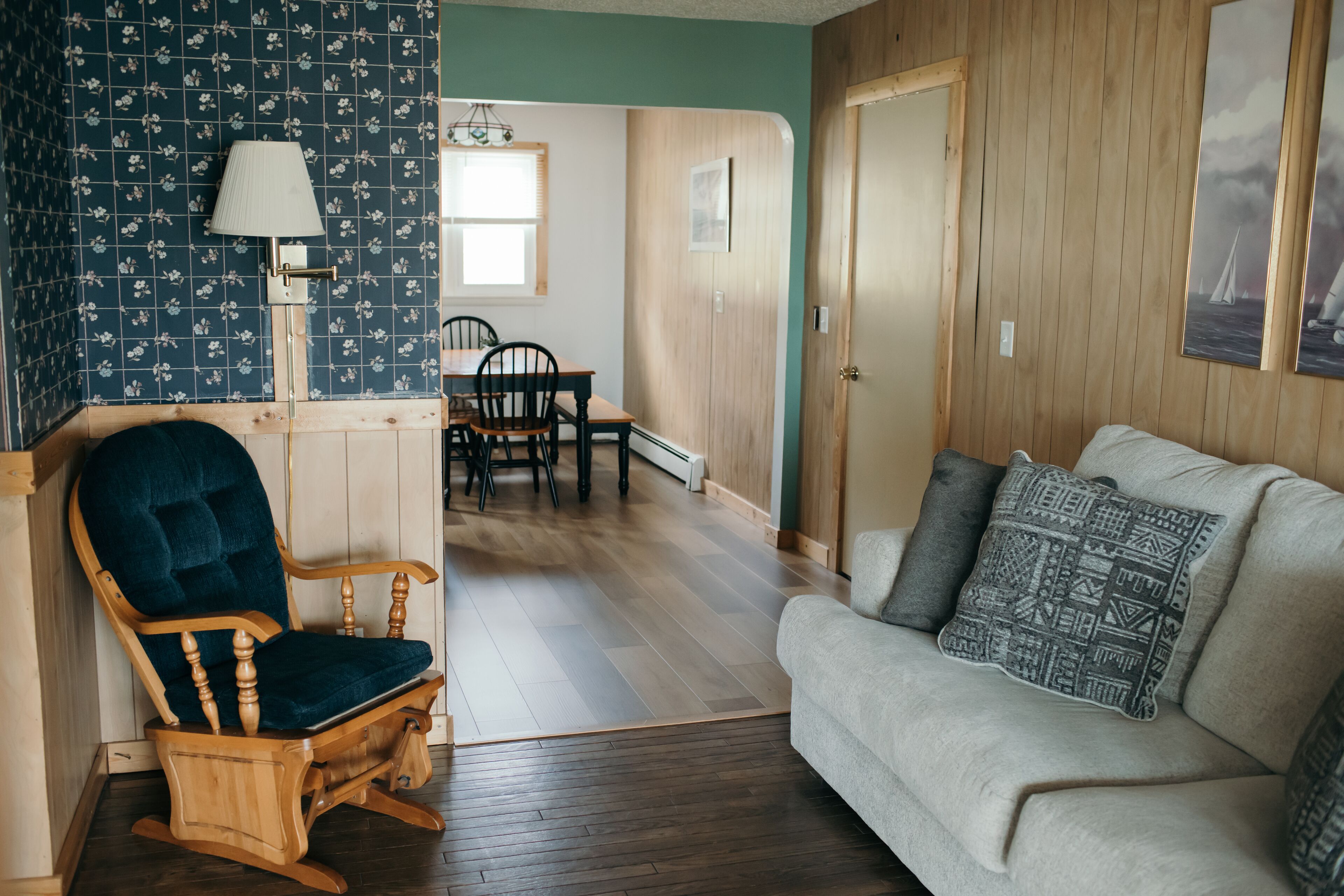 A room with a wooden rocking chair, a grey sofa with patterned cushions, a dining table with chairs, and a wooden door.