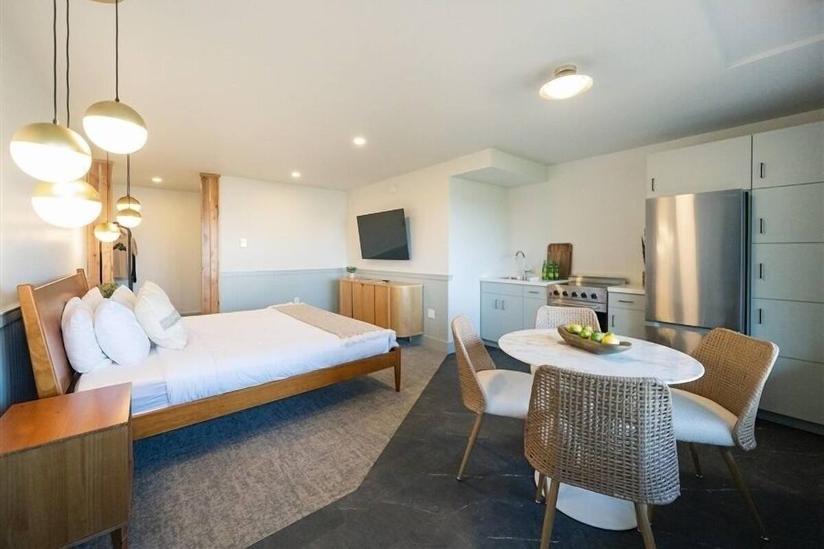A modern hotel room with a large bed, a dining area with wicker chairs, a kitchen with a refrigerator, and a flat-screen TV.