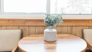 A round wooden table with a white vase and green plant in the center, placed in front of a window with wooden paneled walls and two cushioned benches. - Seagull Bay Motel (Bayfield)