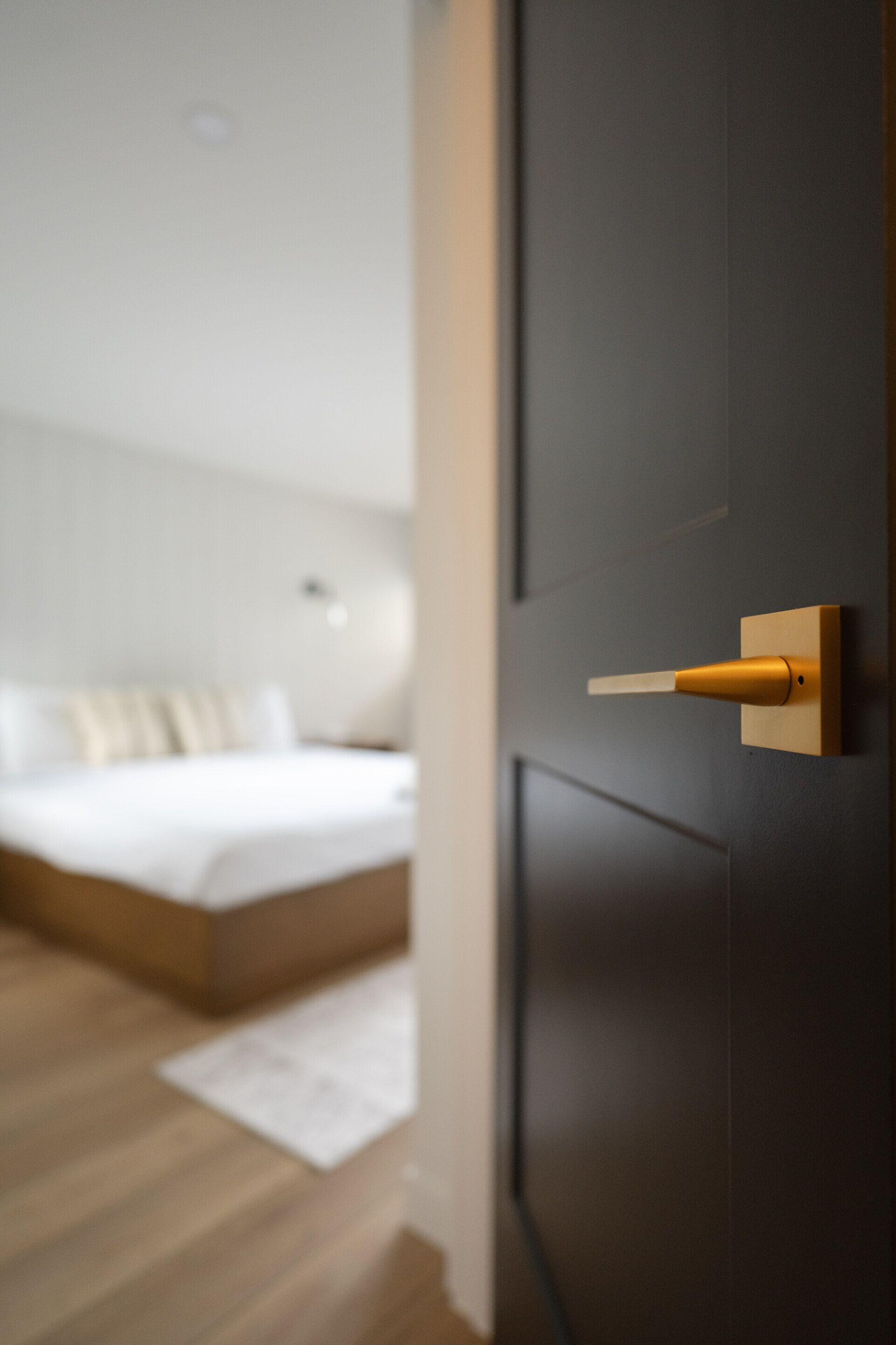 Un dormitorio moderno con diseño minimalista, que presenta una cama con estructura de madera y ropa de cama blanca, una puerta oscura con pomo dorado y un suelo de color claro.