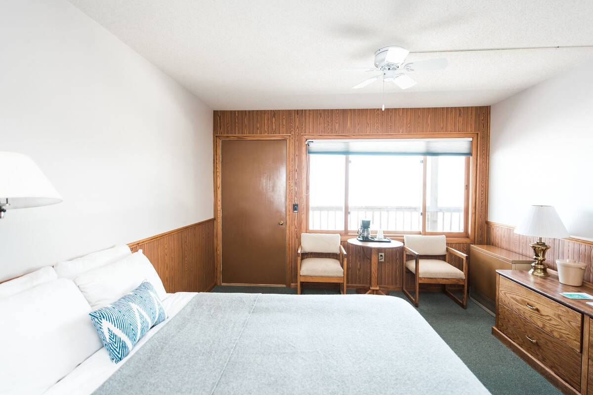 A hotel room with a bed, two chairs, a small table, a desk, and a window with a view.