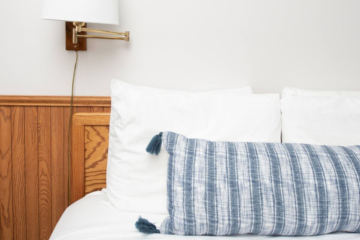 A neatly made bed with white linens and a blue striped pillow, a wall-mounted lamp, and a wooden headboard.