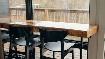 Una terraza de madera con una mesa y sillas, un ventanal grande y vistas a árboles.