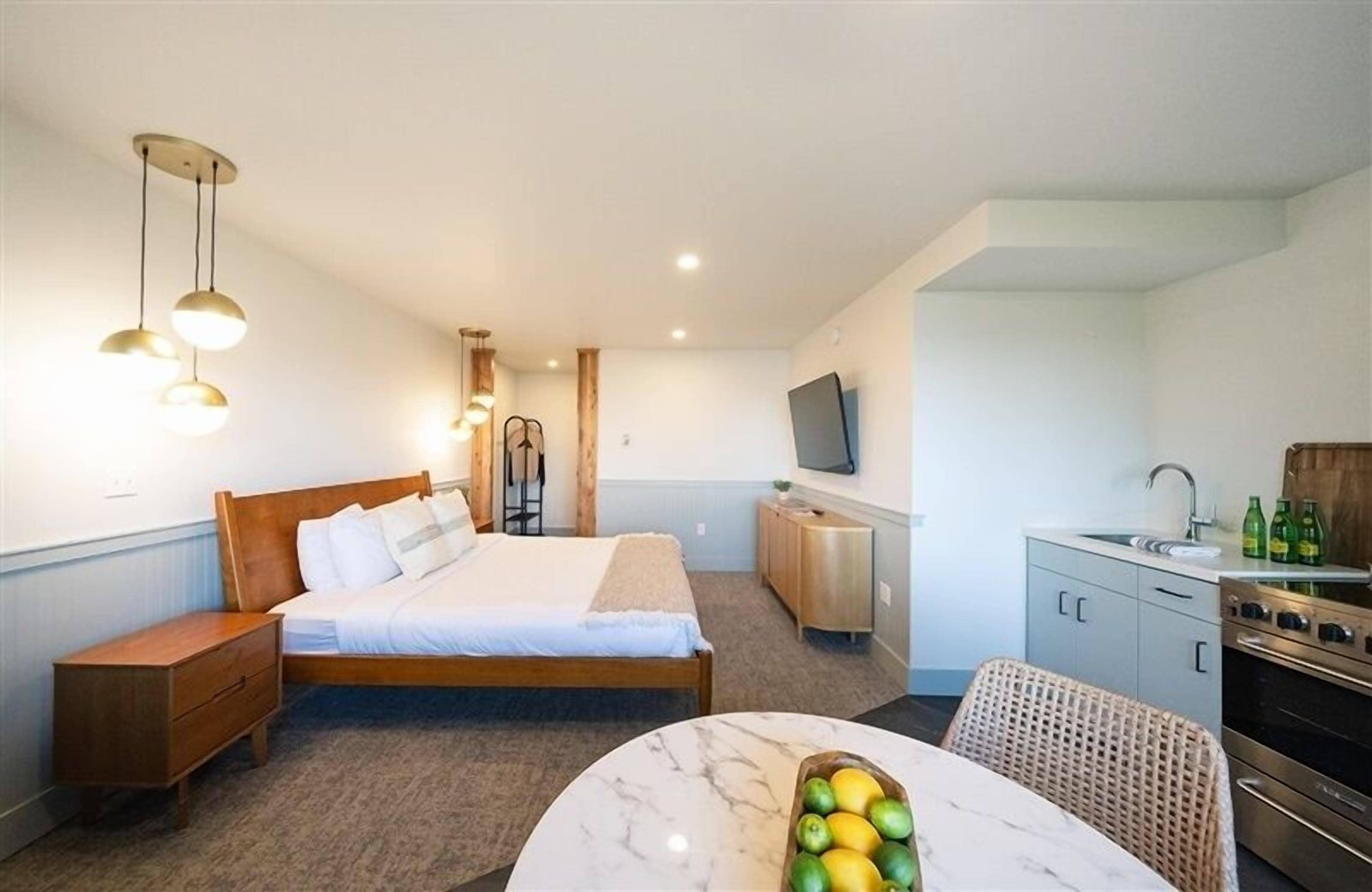A hotel room with a bed, bedside table, a small kitchenette, a dining area with a table and chairs, and a TV mounted on the wall.