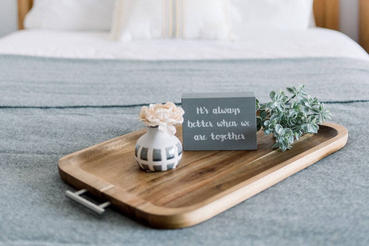 A neatly made bed with white bedding, a small decorative vase, a tray with a card, and a lamp on a bedside table.