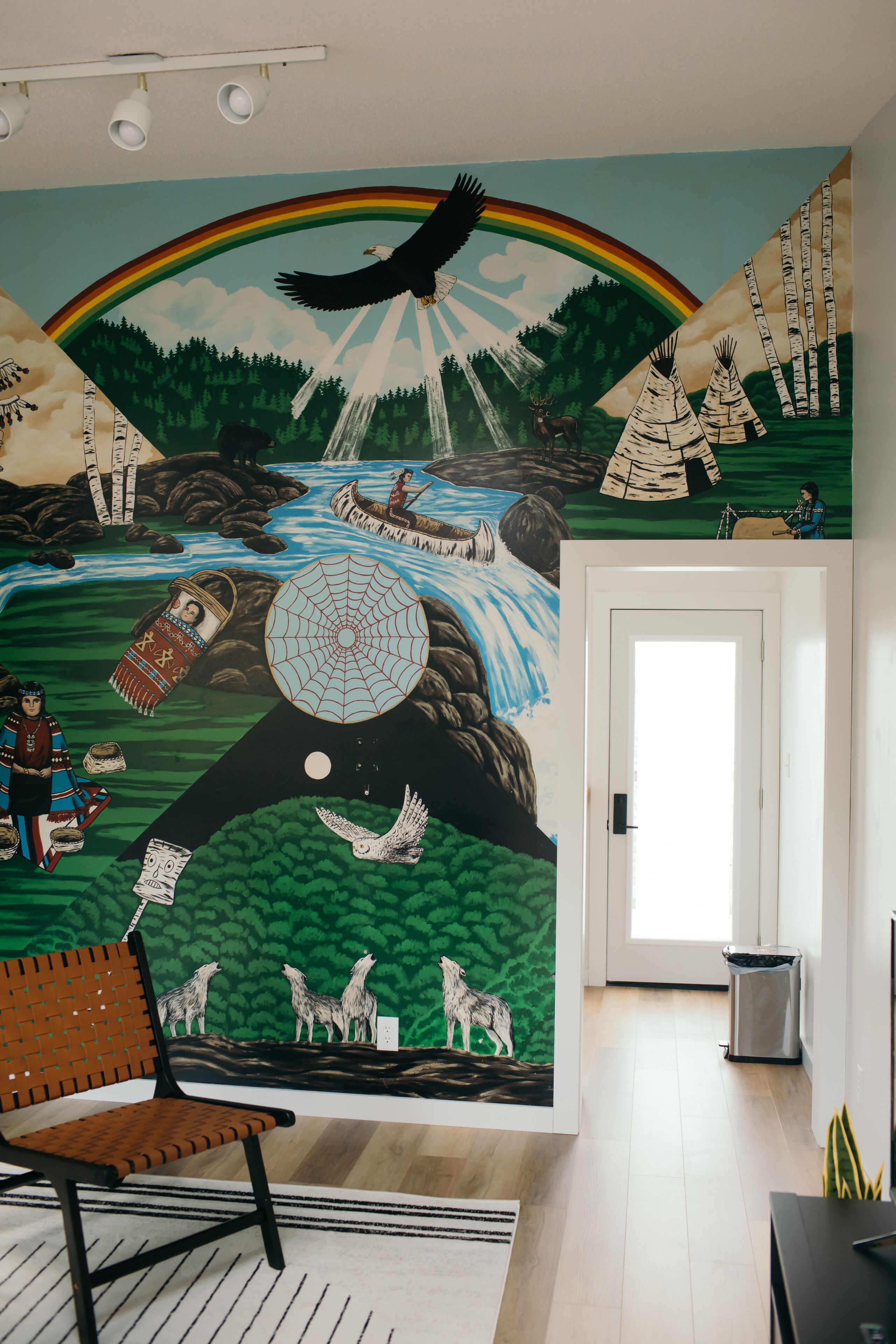 A room with a mural depicting a natural scene with a rainbow, eagle, and totem poles. There is a wooden bench and a door in the room.