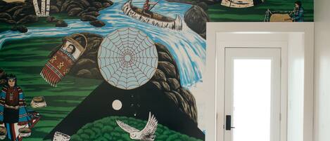 A room with a mural depicting a natural scene with a rainbow, eagle, and totem poles. There is a wooden bench and a door in the room.