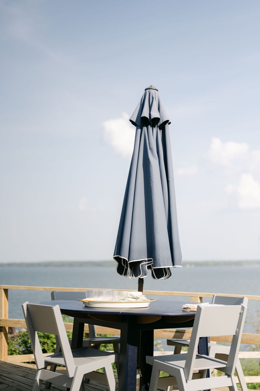 A table with a blue umbrella, two white chairs, and a plate with a napkin on a wooden deck overlooking a body of water.