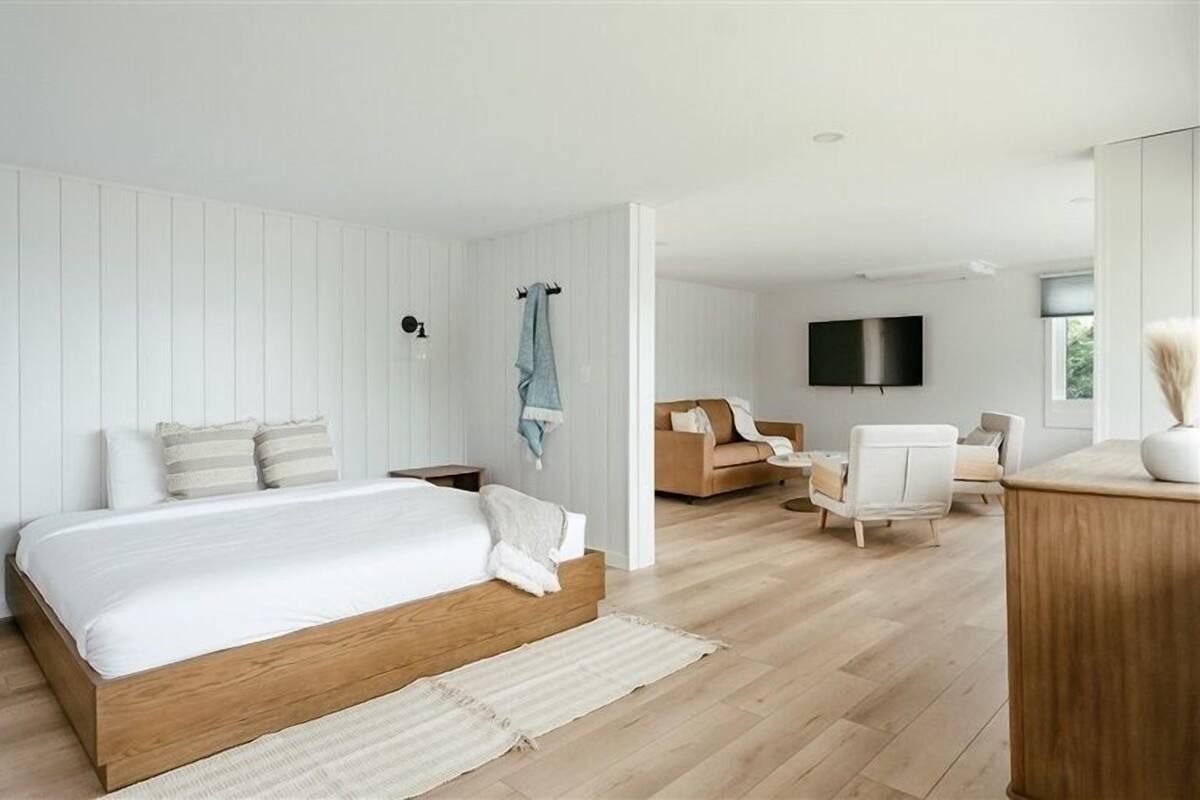A modern bedroom with a wooden bed, a TV mounted on the wall, and a seating area with a sofa and chairs.