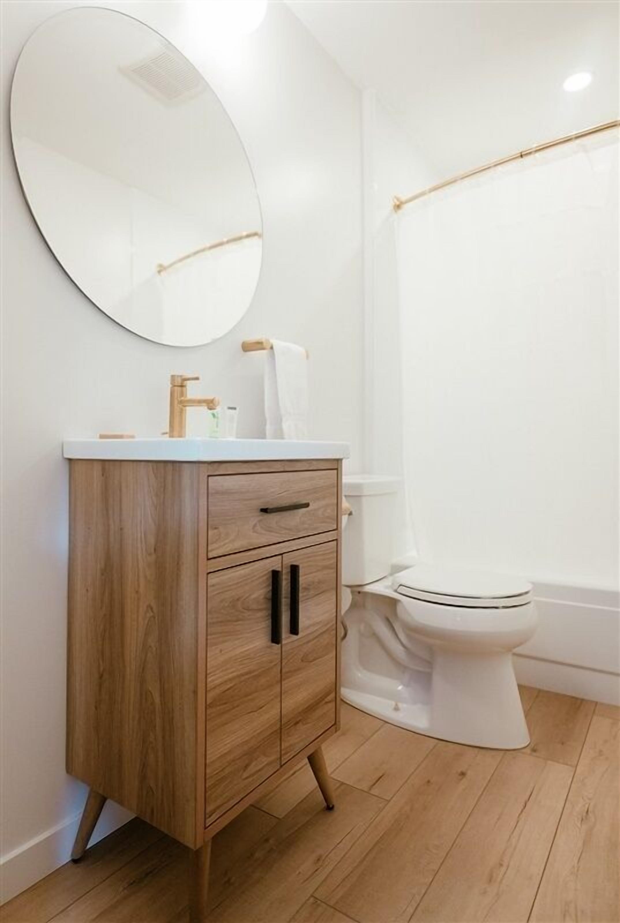 A bathroom with a wooden vanity, a round mirror, a white toilet, and a shower curtain rod.