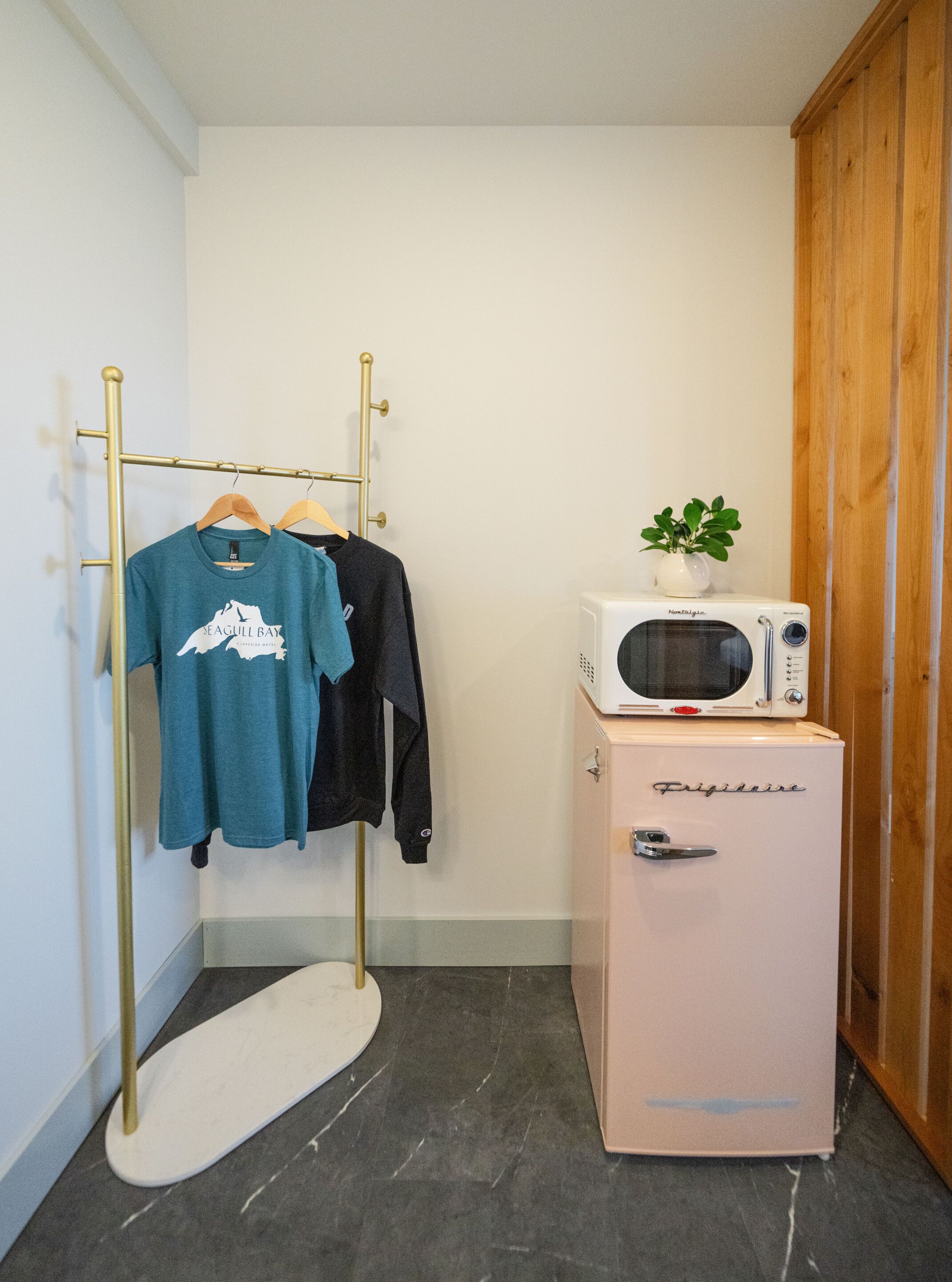 A compact room with a white wardrobe, a small white refrigerator, a microwave, and a clothing rack with two shirts.