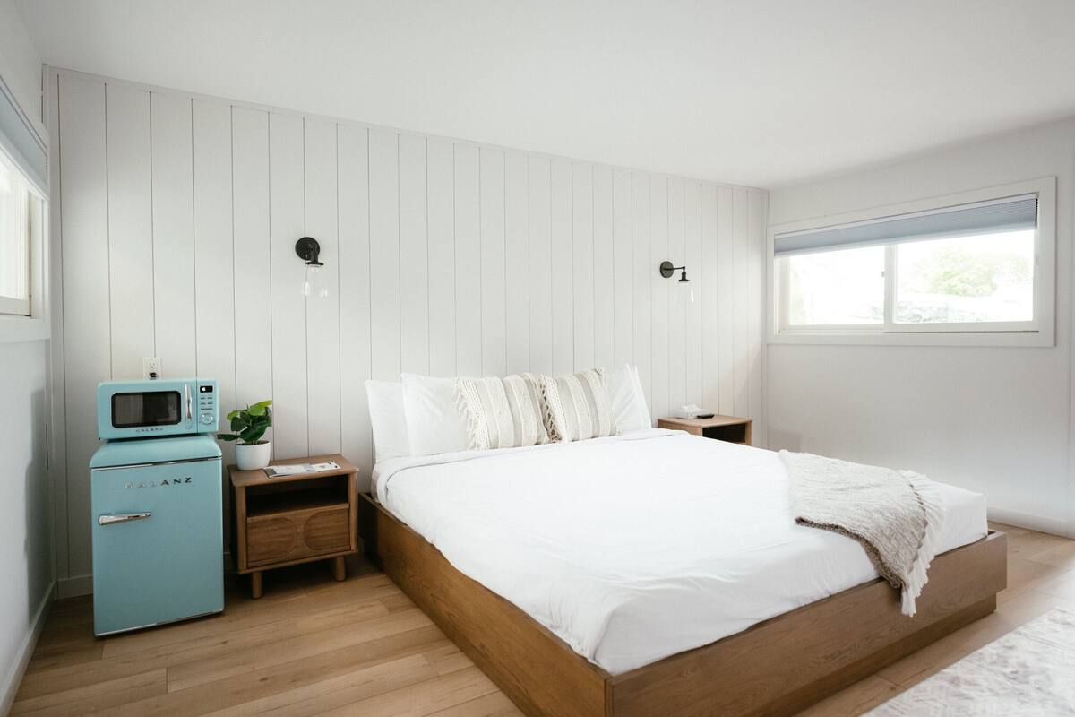 A bedroom with a wooden bed, a small teal-colored microwave, a nightstand, and a window.