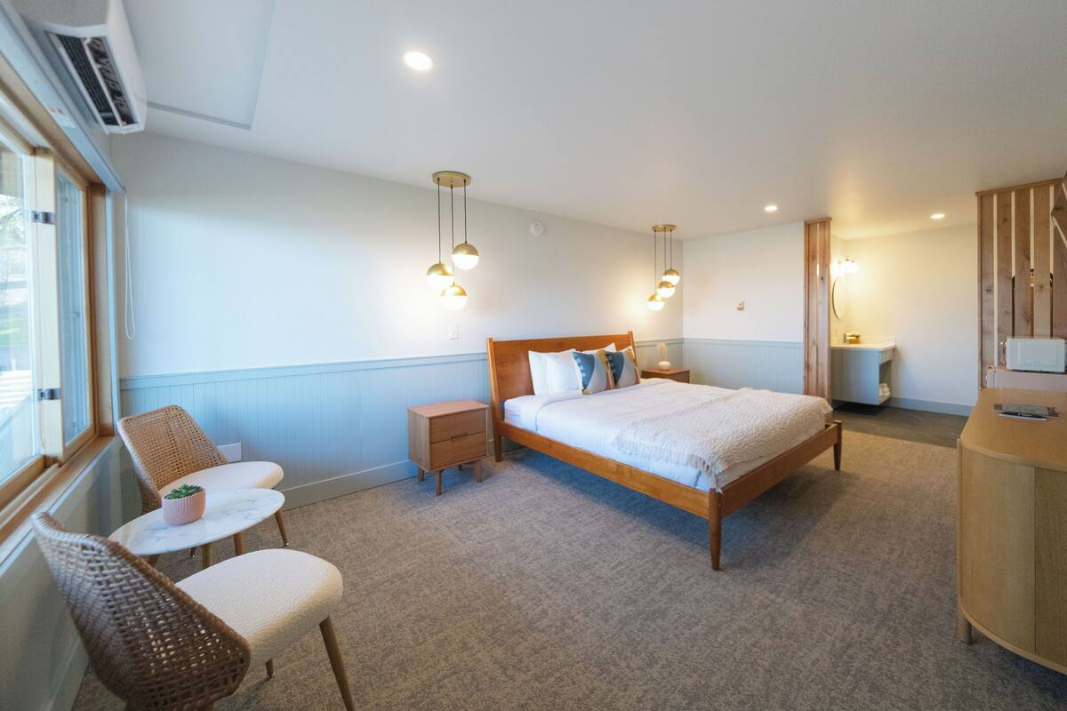 A bedroom with a wooden bed, bedside tables, a small round table with chairs, a window, and a built-in TV.