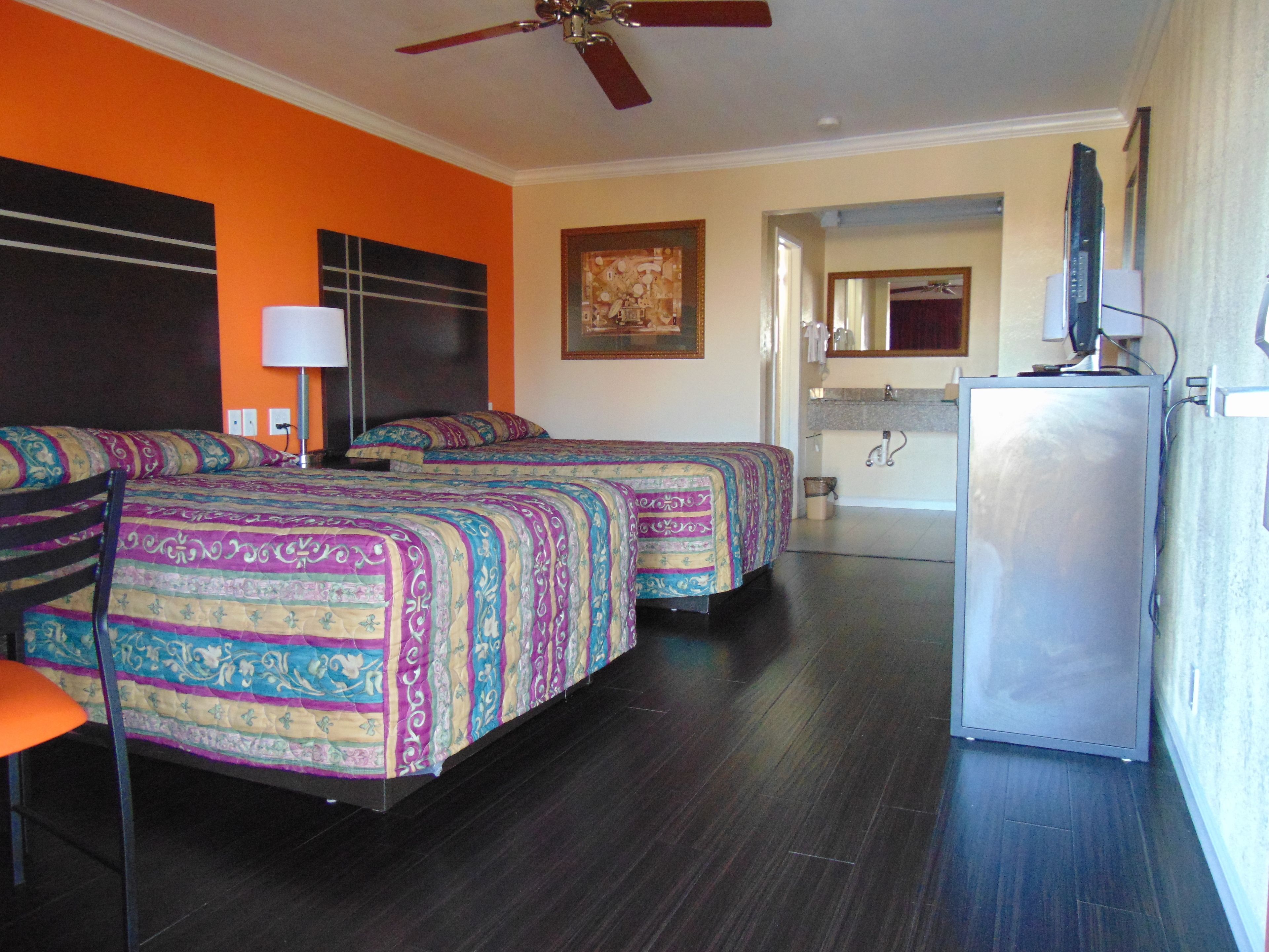 A hotel room with two beds, a desk, a chair, a ceiling fan, a television, and a bathroom visible in the background.