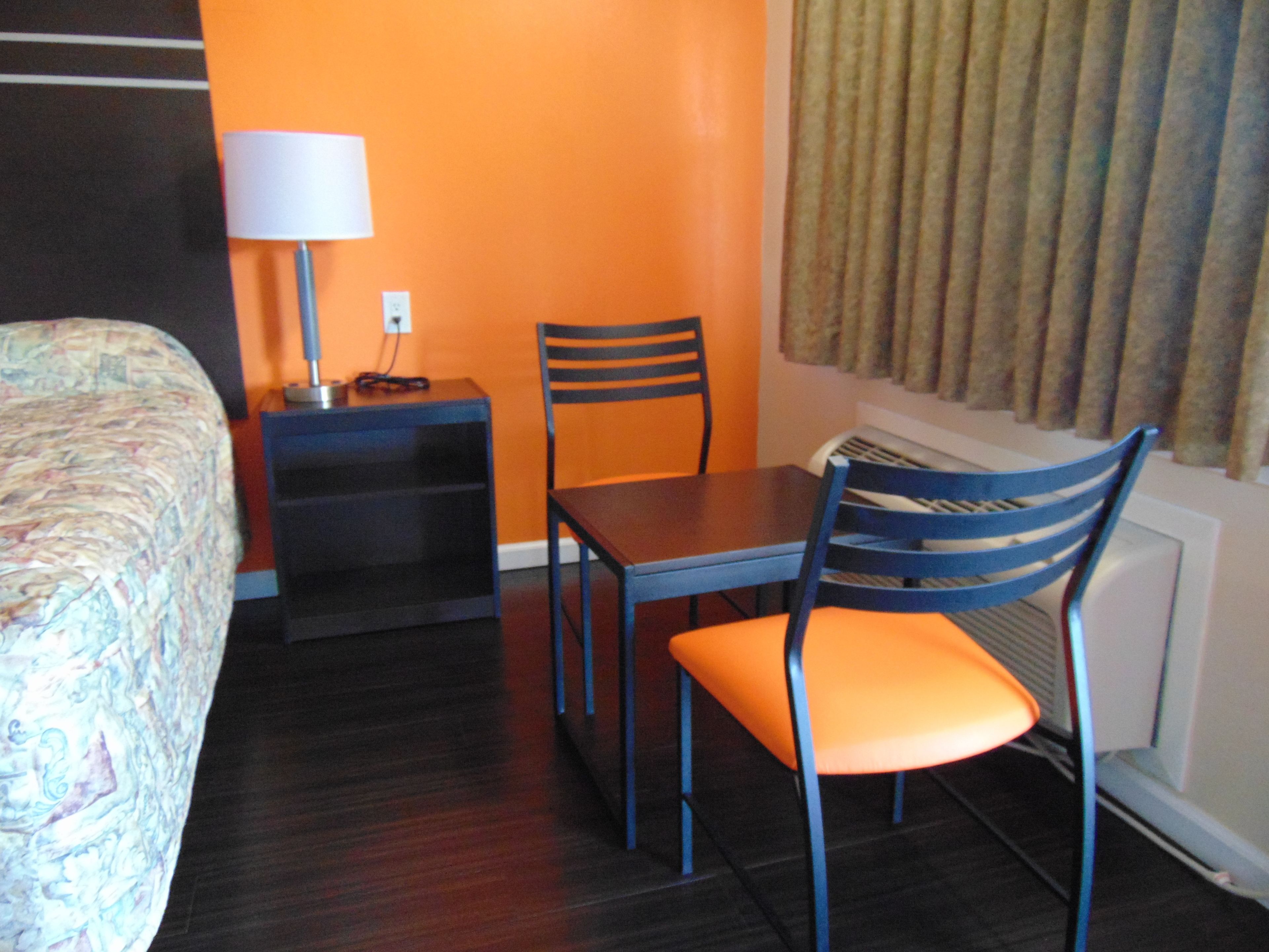 A hotel room with a bed, a desk with a lamp, an orange chair, and a window with curtains.