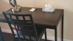 Hotellihuoneessa on tumma puinen työpöytä, tuoli, lamppu, paperipyyhekotelo ja peili seinällä.