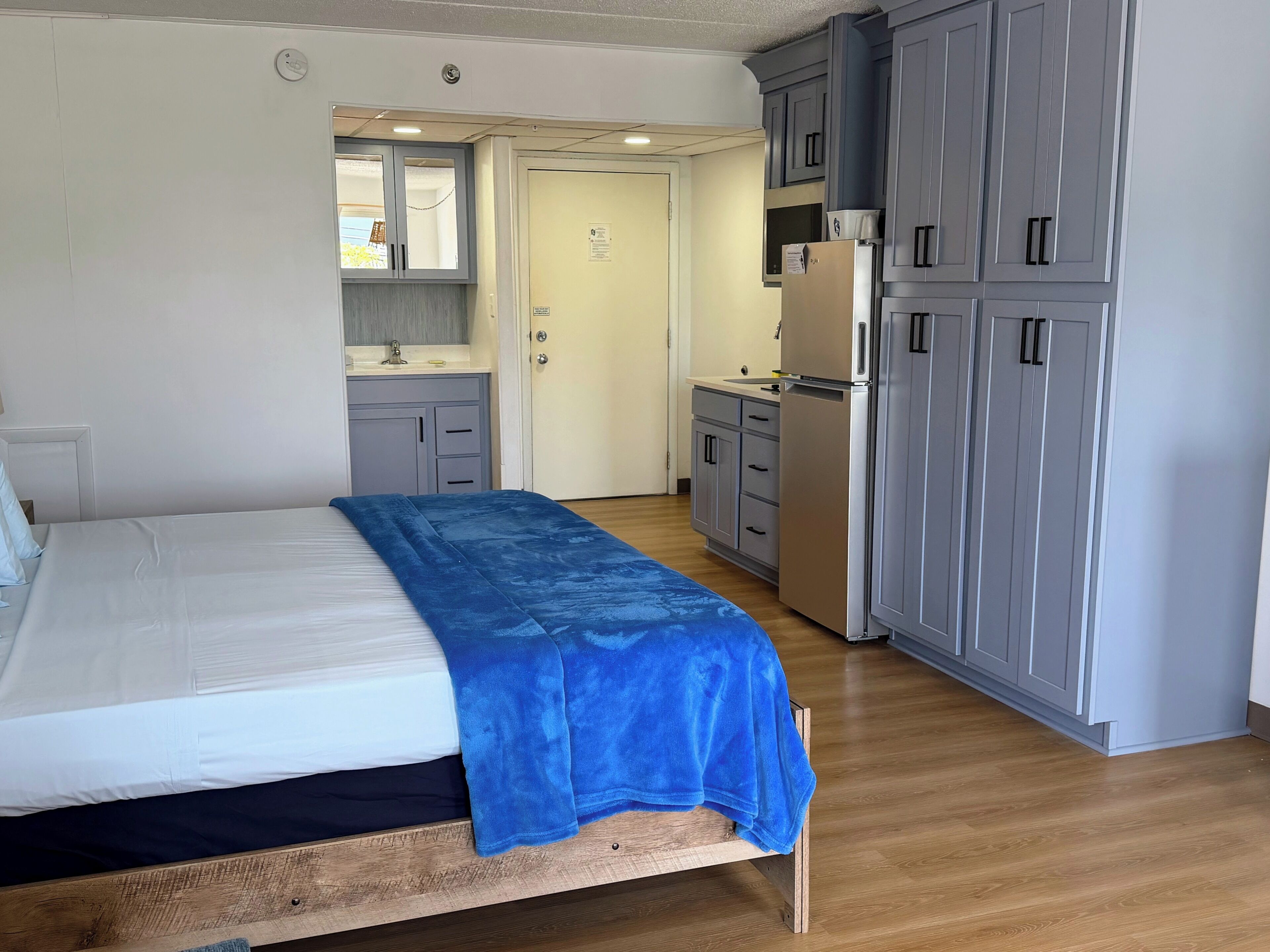 A bedroom with a bed, a blue blanket, a wooden headboard, a small kitchenette with a fridge, and grey cabinets.