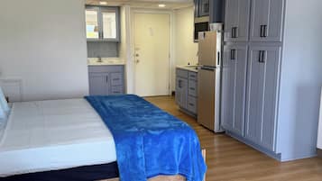 A bedroom with a bed, a blue blanket, a wooden headboard, a small kitchenette with a fridge, and grey cabinets.