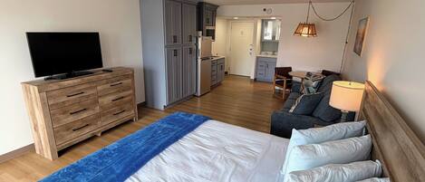 A bedroom with a bed, a TV, a dresser, a sofa, and a kitchen in the background.