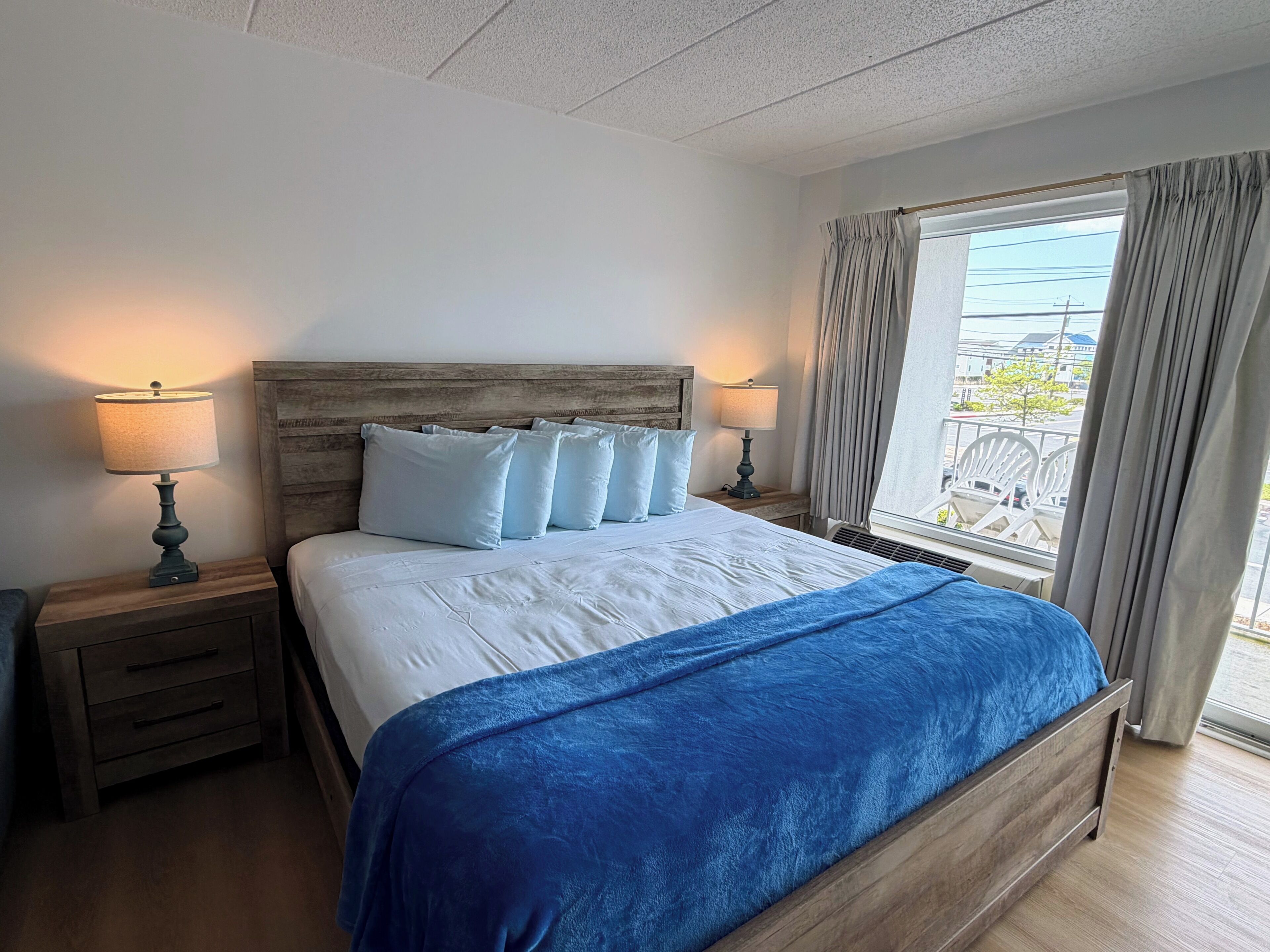 A hotel room with a wooden headboard, a large bed with white bedding and a blue throw, two bedside tables with lamps, and a window with curtains.