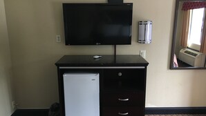 A room with a flat-screen TV mounted on a dark dresser, a small white refrigerator, a wall-mounted air conditioner, and a mirror reflecting part of the room. - Del Amo Inn (Torrance)
