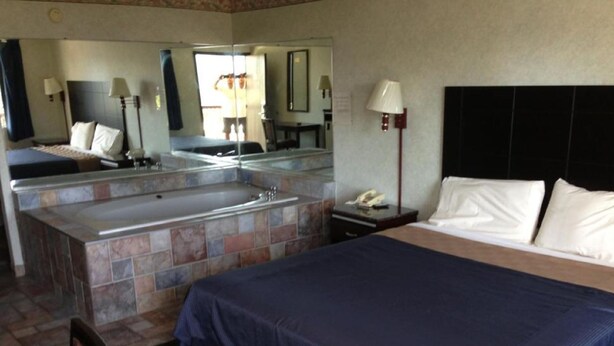 A hotel room with a bed, a large mirror, a bathroom with a bathtub, and a nightstand with a lamp. - Del Amo Inn (Torrance)