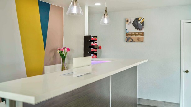 A reception desk with a modern design, featuring a white counter and a colorful wall. There are two pendant lights above the desk, and a small vase with flowers on the counter. - Studio 6 Suites Albany, OR (Albany)