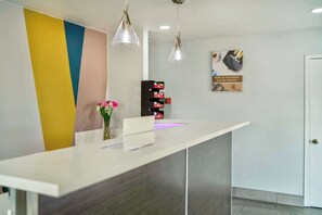 A reception desk with a modern design, featuring a white counter and a colorful wall. There are two pendant lights above the desk, and a small vase with flowers on the counter. - Studio 6 Suites Albany, OR (Albany)