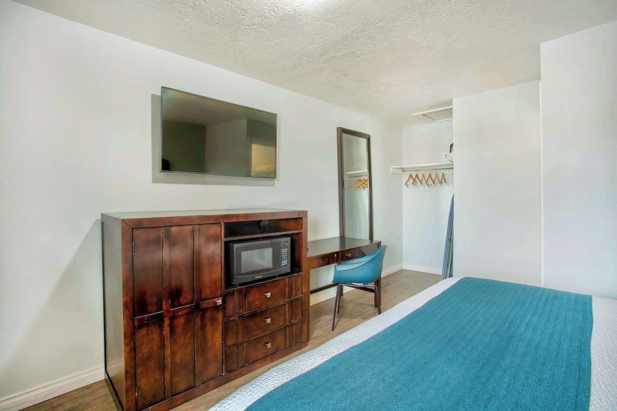 A bedroom with a bed, a wooden dresser with a microwave, a blue chair, and a mirror.