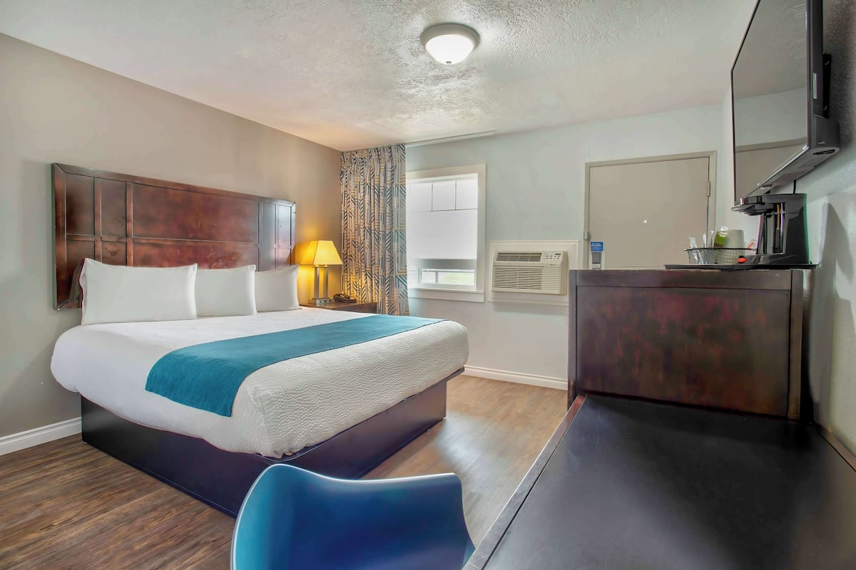 A hotel room with a bed, a desk, a chair, a lamp, a mirror, and an air conditioning unit.