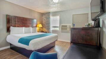 A hotel room with a bed, a desk, a chair, a lamp, a mirror, and an air conditioning unit.