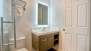 A bathroom with a toilet, a sink, a mirror, and a shower area. - Studio 6 Suites Albany, OR (Albany)