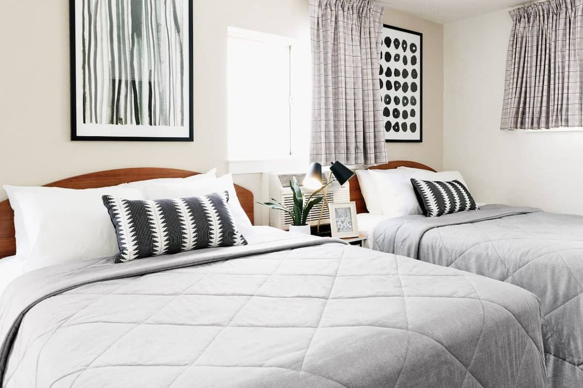 A hotel room with two beds, each with a grey quilted duvet and patterned pillows. There are framed pictures on the nightstands, a lamp, and a window with curtains.