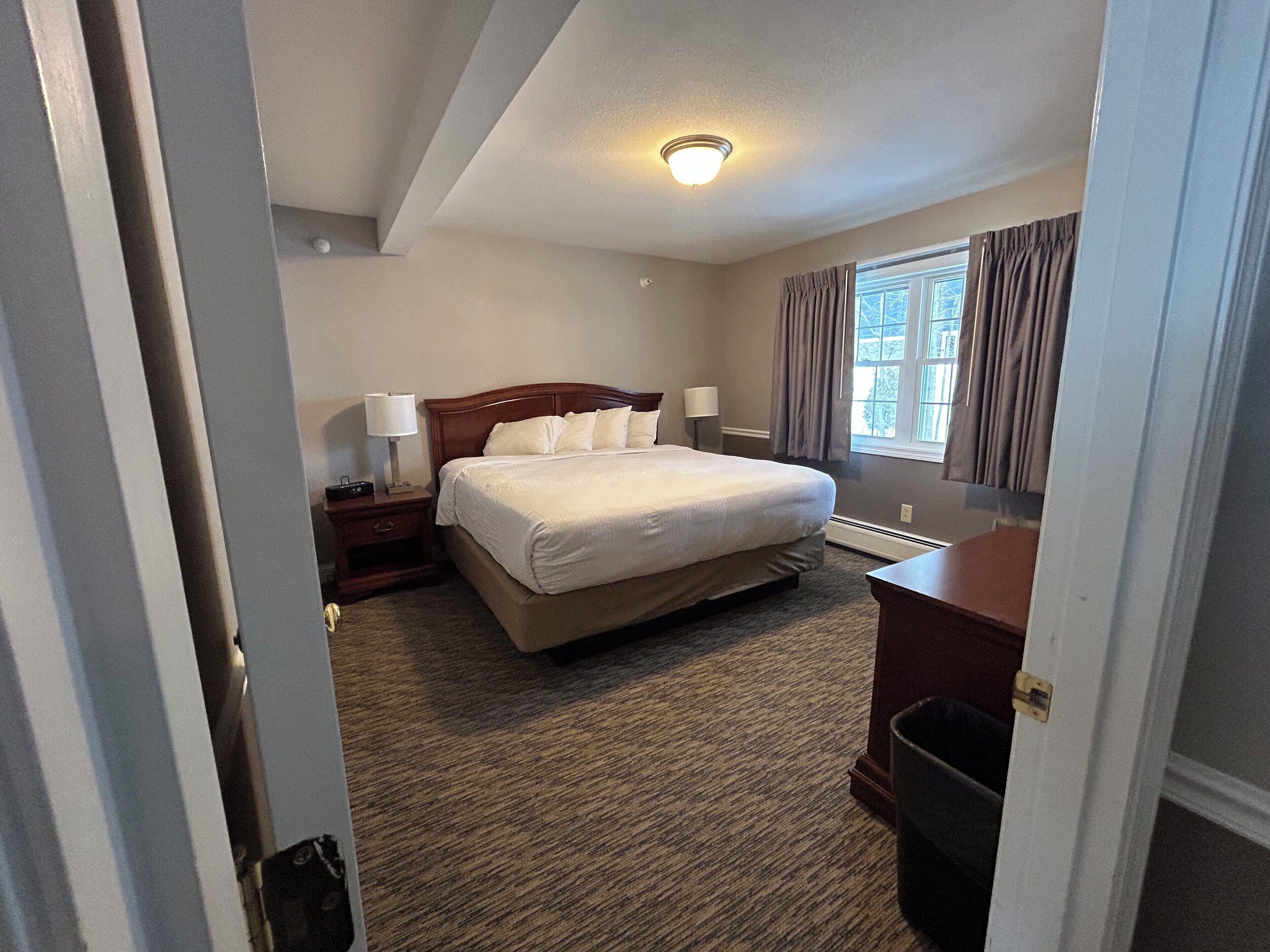 Une chambre d’hôtel avec un grand lit, une table de chevet avec une lampe, une commode et une fenêtre avec des rideaux.