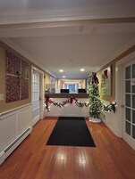 Un pasillo con piso de madera, un árbol de Navidad decorado con luces y guirnaldas, y una puerta a la derecha.